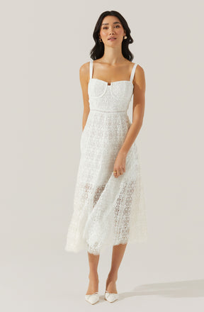 A woman is wearing a sleeveless, white lace dress with wide shoulder straps. The dress features a fitted bodice with decorative lace detailing around the neckline and a long, flowing skirt made of sheer lace fabric that extends to her mid-calf. She is accessorized with gold earrings and a gold ring, and is wearing white pointed-toe shoes with bow details.