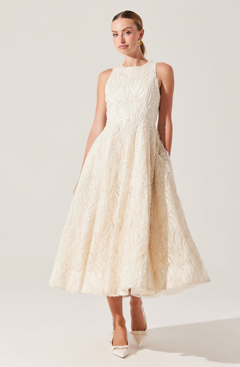 A woman is wearing a sleeveless, high-neck, full-length dress in an off-white or cream color. The dress features textured, intricate patterns throughout, giving it a luxurious and elegant appearance. She pairs the dress with pointed-toe heels and gold earrings.