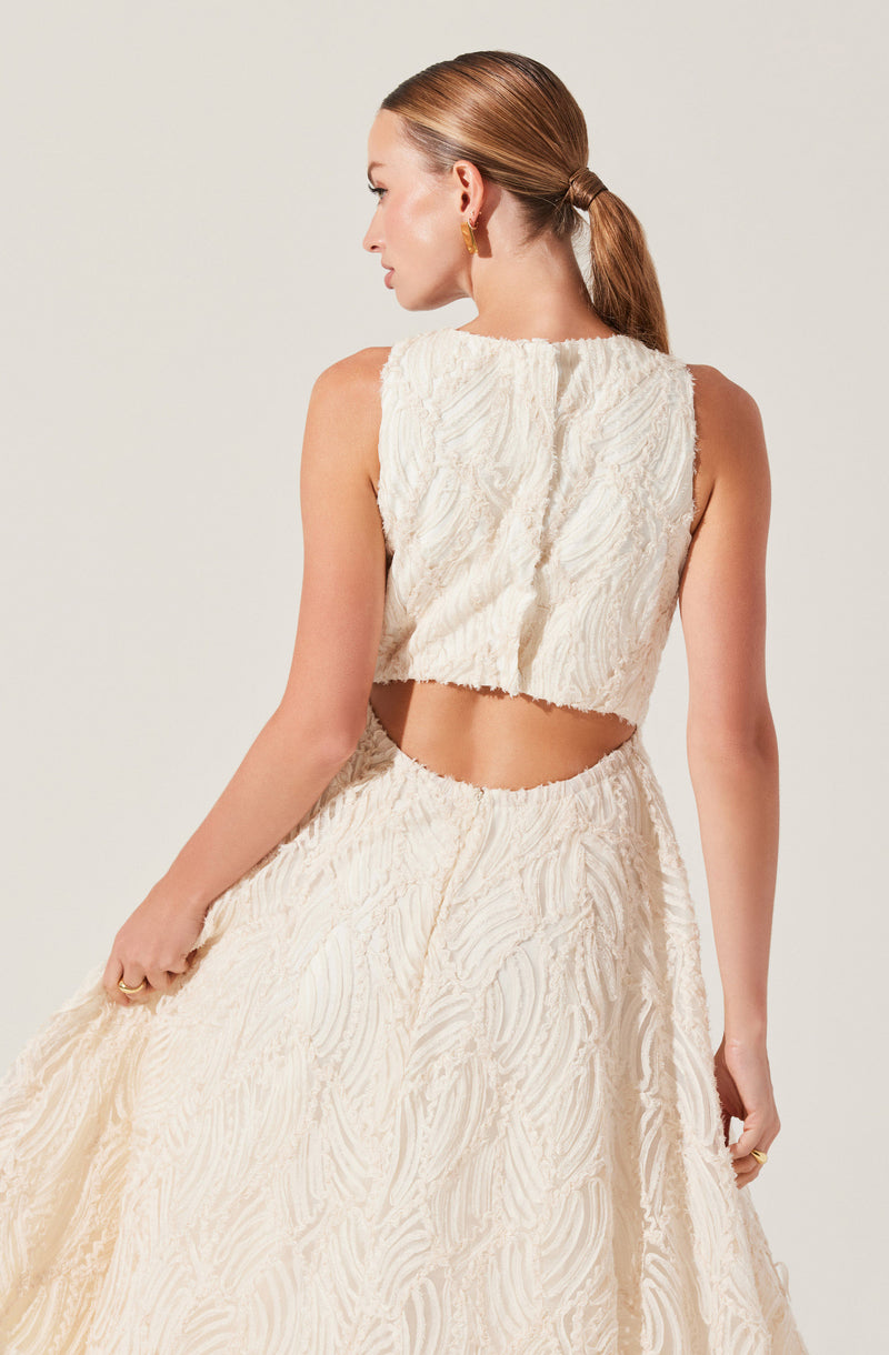 A woman is wearing a sleeveless, off-white outfit made of textured fabric with a swirling pattern. The top is cropped and has a rounded neckline, revealing her midriff. The matching skirt is high-waisted and extends to the floor, creating an elegant and cohesive look. She has her hair tied back in a low ponytail and is accessorized with gold earrings and rings.