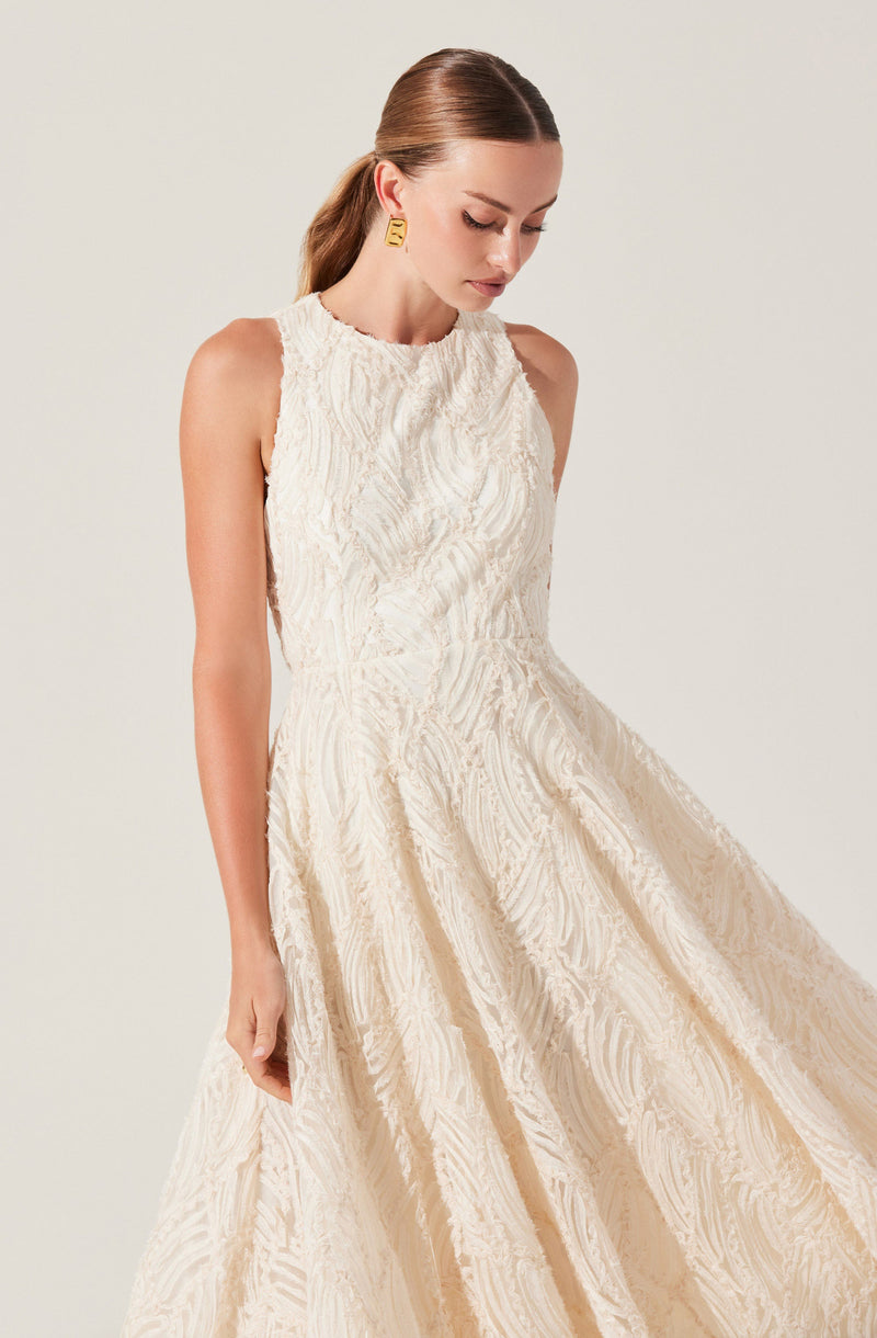 A woman is wearing a sleeveless, high-neck, full-length dress made of textured, cream-colored fabric with a subtle pattern. She has her hair pulled back and is accessorizing with a pair of rectangular, gold earrings.