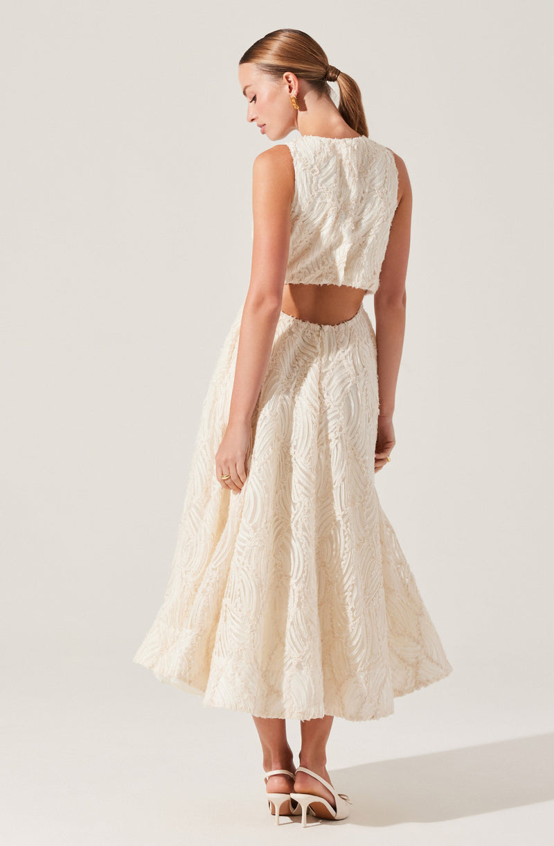 A woman is wearing a two-piece cream-colored textured outfit. The top is a sleeveless, cropped piece with a high neckline. The skirt is high-waisted, full-length, and features the same textured fabric as the top. She has her hair tied back in a low ponytail and is wearing gold hoop earrings. She is also wearing white high-heeled sandals.