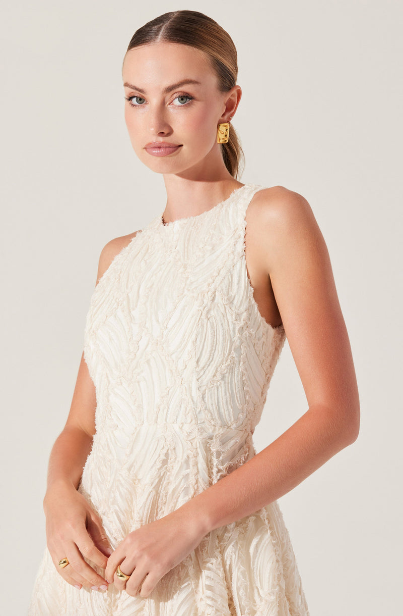 A woman is wearing a sleeveless, textured, cream-colored dress with a high neckline. She has her hair pulled back and is accessorized with large, gold, geometric earrings and rings on her fingers.