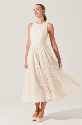A woman is wearing a sleeveless, full-length cream-colored dress with a textured, patterned fabric. The dress features a fitted bodice and a flowing, slightly flared skirt. She is also wearing cream-colored heels with a bow detail at the toe.