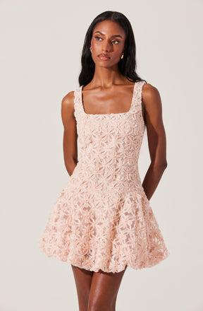 A woman is wearing a sleeveless, peach-colored dress with a floral lace overlay. The dress has a square neckline and features intricate flower patterns throughout. The skirt is slightly flared, creating a A-line silhouette.