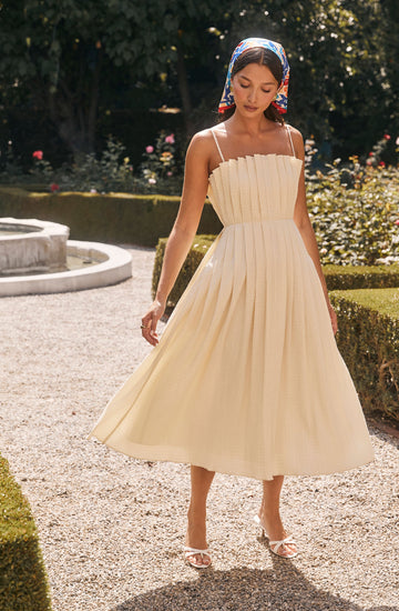 A woman is wearing a cream-colored, pleated midi dress with thin spaghetti straps. She has a multicolored headscarf tied around her head. She is also wearing white high-heeled sandals with an open toe. The background features a garden with greenery, flowers, and a small round fountain.