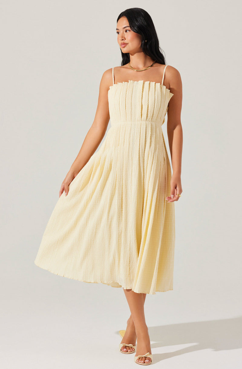 A woman is wearing a sleeveless, light yellow dress with thin spaghetti straps. The dress has a textured, pleated fabric that creates a ruffled effect around the neckline and flows down to a midi length. She is also wearing open-toe, high-heeled sandals in a matching light yellow color.