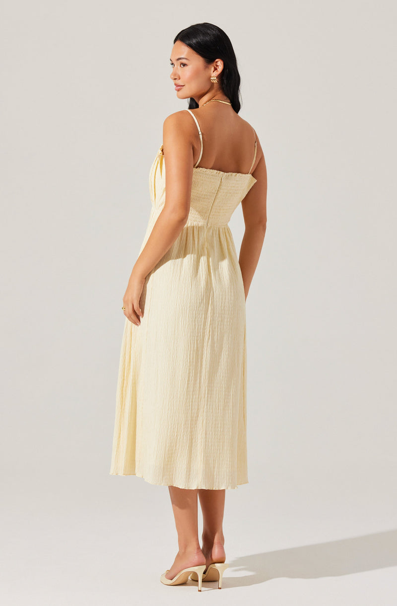 A woman is wearing a light yellow, sleeveless, midi-length dress with thin spaghetti straps and a gathered, elasticized waist. The fabric has a slightly textured, crinkled appearance. She is paired with nude high-heeled sandals.