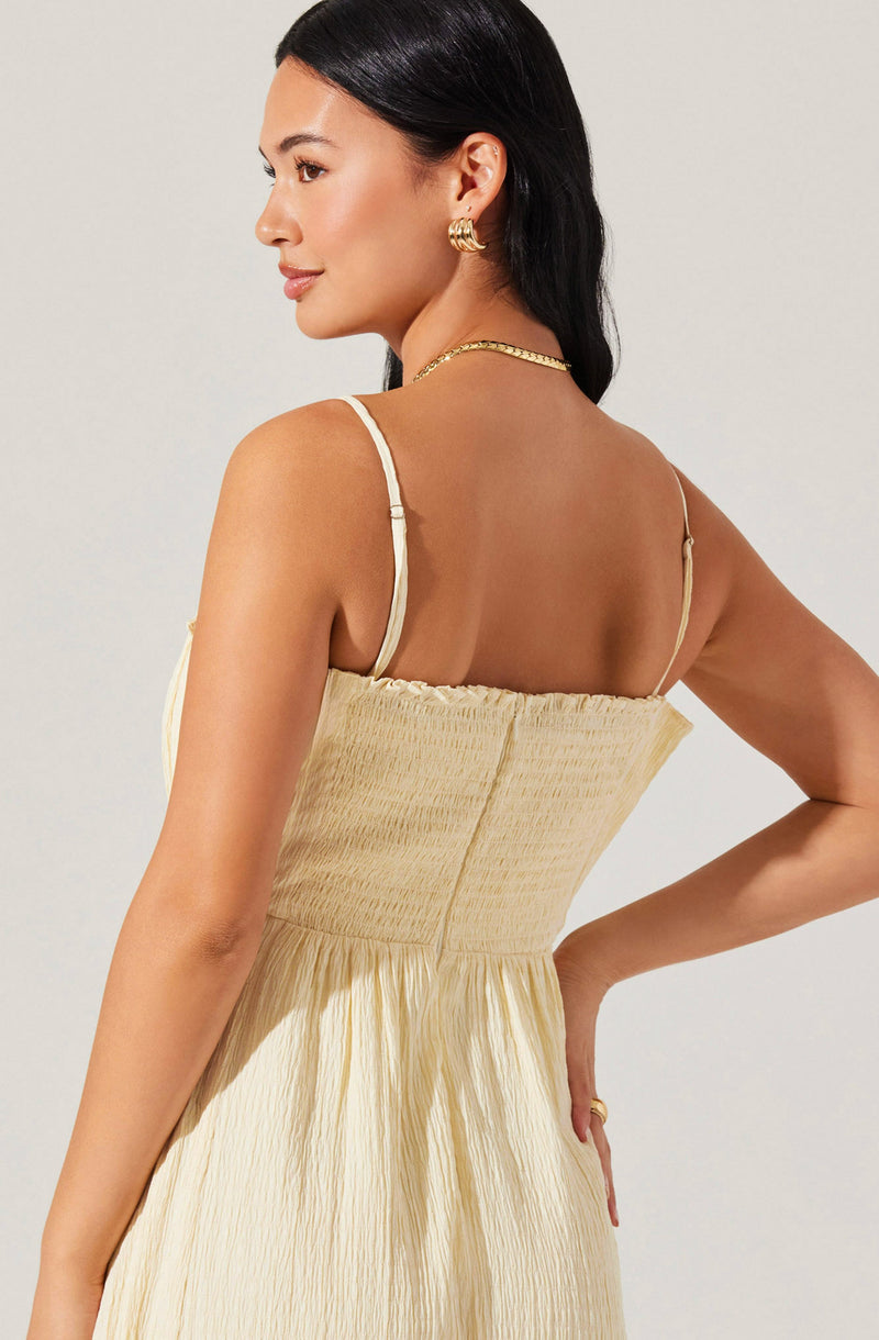 A woman is wearing a light yellow, sleeveless dress with thin spaghetti straps. The dress features a fitted, smocked bodice and a flowing, crinkled fabric skirt. She accessorizes with a gold chain necklace, gold hoop earrings, and a gold ring. Her black hair is styled straight and parted on the side.