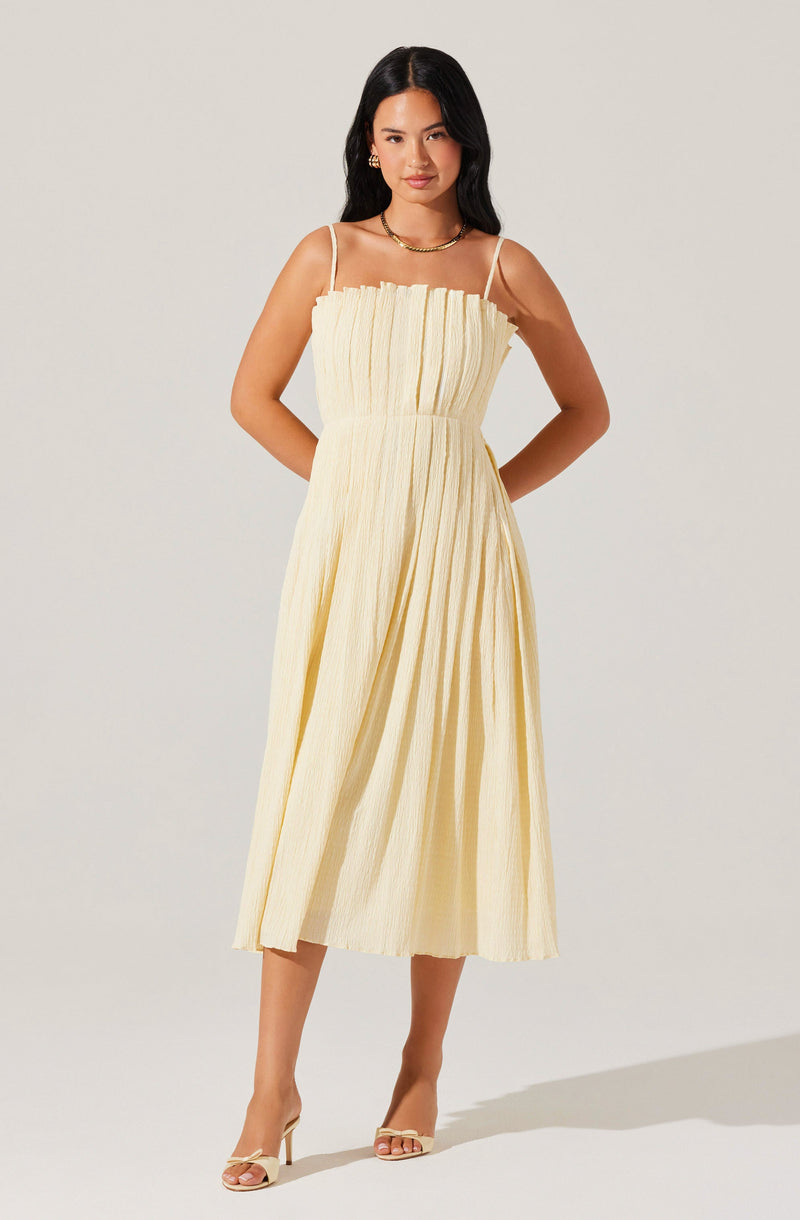 A woman is wearing a light yellow, sleeveless, ankle-length dress with thin spaghetti straps. The dress features a gathered, pleated bodice and a full, flowing skirt with subtle vertical stripes. She has accessorized with gold jewelry, including hoop earrings and a chain necklace, and is wearing open-toed, beige high-heel sandals.