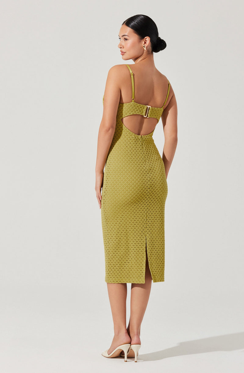 A woman wearing a fitted, sleeveless dress in a green geometric pattern. The dress features adjustable shoulder straps, a wide waistband with a metallic buckle, and a cutout detail at the back with a small slit at the hem. She is also wearing light-colored high-heeled shoes.
