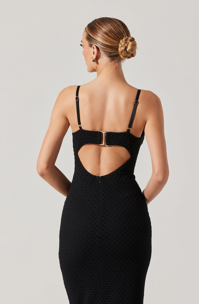 A woman is wearing a fitted black dress with a textured fabric. The dress features thin black shoulder straps and a distinctive cut-out detail at the back, held together by a gold-toned clasp. The dress extends to below the hips, creating a sleek silhouette.