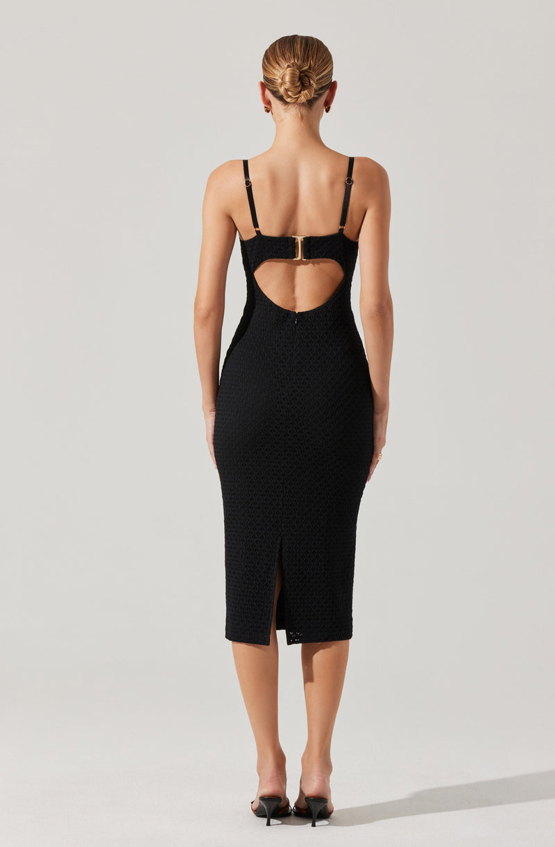 A woman is wearing a fitted black dress with thin shoulder straps. The dress features a large round cutout on the back, secured with a gold buckle at the top. The hemline of the dress falls below the knees with a small slit at the back. The fabric appears textured, and she is wearing black high-heeled shoes.