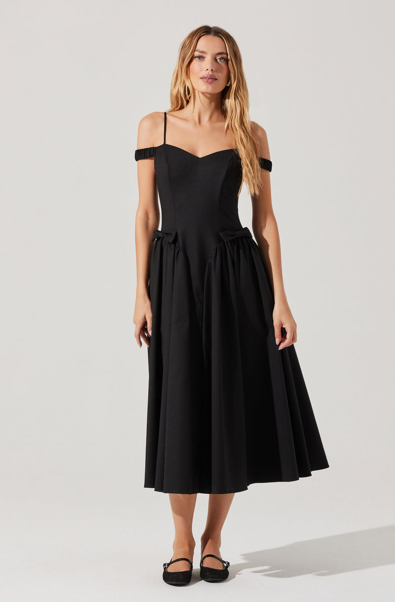 A woman is wearing a black dress with spaghetti straps and off-the-shoulder ruffled sleeves. The bodice is fitted, and the skirt is full and gathered, extending to mid-calf length. She is also wearing black shoes with ankle straps.