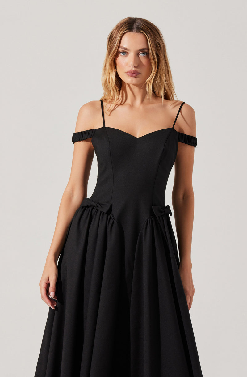 A woman is wearing a black gown with thin spaghetti straps and off-the-shoulder elasticized ruffle details. The dress features a fitted bodice with a slight sweetheart neckline, and the skirt has gathered fabric at the waist with decorative bows, creating a full, flowing silhouette.