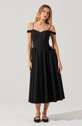 A woman is wearing a black dress with thin spaghetti straps and off-the-shoulder ruffle details around the upper arms. The bodice is fitted, and the skirt is voluminous with pleats, reaching mid-calf length. She is also wearing black shoes with ankle straps.