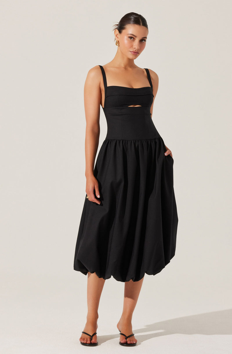 A woman is wearing a black sleeveless dress with wide shoulder straps, a fitted bodice with horizontal seam detailing and a small cutout below the chest, and a voluminous, gathered skirt that falls below the knees. She is also wearing black high-heeled sandals and gold earrings.