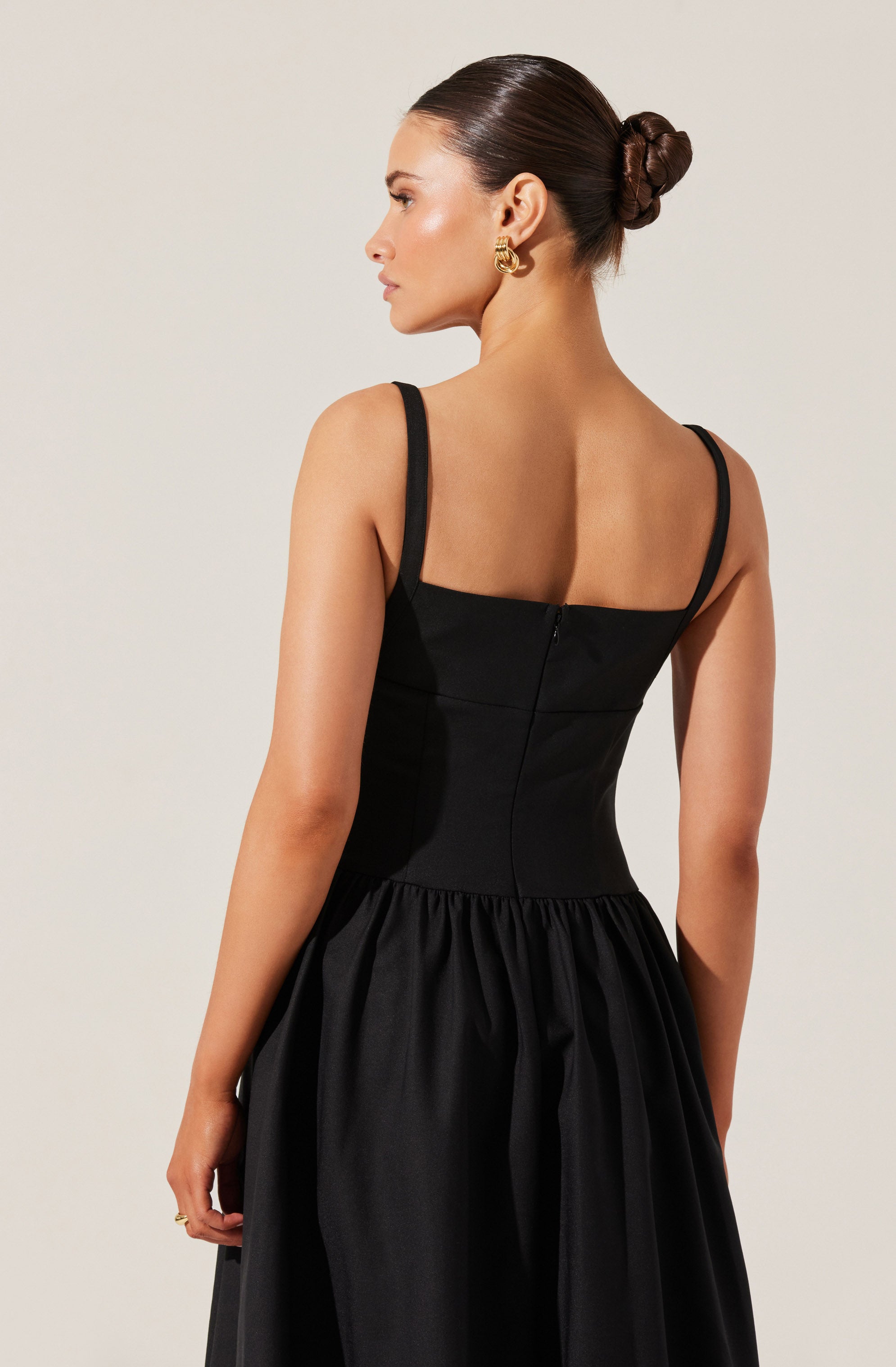 A woman is wearing a sleeveless black dress with thin straps and a fitted bodice. The dress features a gathered skirt that falls from the waist. She has her dark hair styled in a sleek, low bun and is wearing gold hoop earrings.