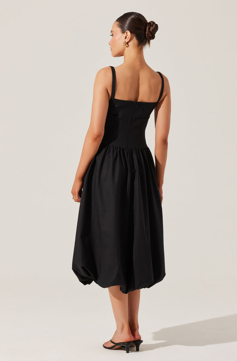 A woman is wearing a black sleeveless dress with thin straps. The dress has a fitted bodice and a gathered, voluminous skirt that reaches below the knees with a bubble hem. She is also wearing black open-toe heels and large gold earrings, with her hair styled in a neat bun.
