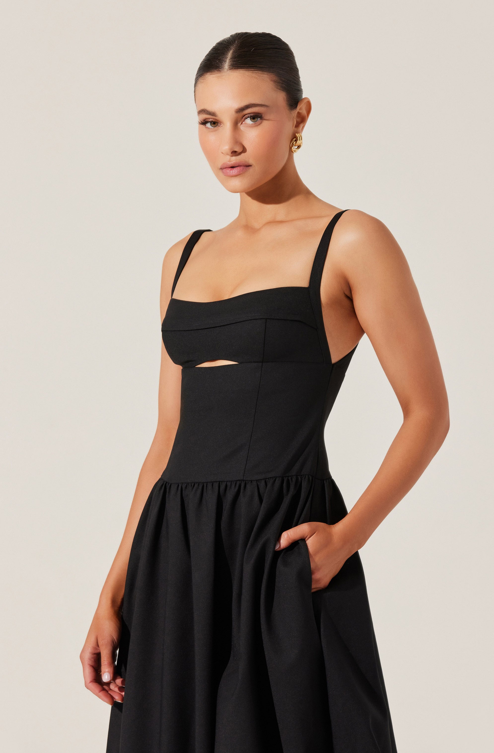 A woman is wearing a sleeveless black dress with wide shoulder straps. The dress has a fitted bodice with a horizontal band across the chest and a small cutout just below the band. The skirt is gathered at the waist, creating a full, voluminous look, with one hand in a pocket. She is also wearing gold earrings and has her dark hair styled in a sleek, center-parted bun.
