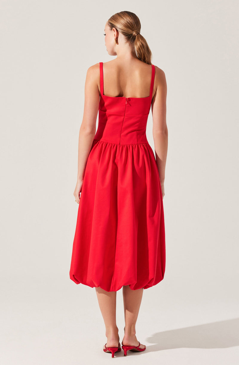 A woman is wearing a red dress with a fitted bodice and wide shoulder straps. The skirt is voluminous with gathered fabric and a balloon-style hem that falls below the knees. She is also wearing matching red heeled sandals.