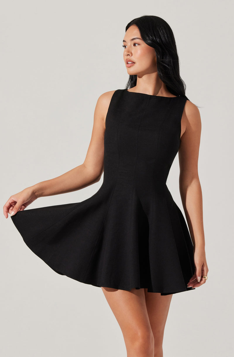 A woman is wearing a sleeveless, black dress with a fitted bodice and a flared, pleated skirt that extends to mid-thigh. The dress has a high, rounded neckline.