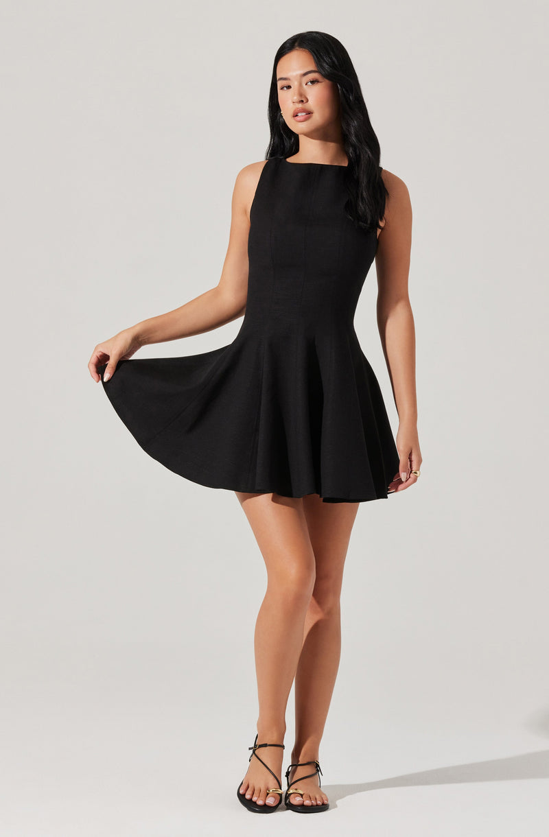A woman is wearing a sleeveless, fitted black dress with a round neckline. The dress has a flared skirt that she is holding out slightly with one hand. She is also wearing black strappy sandals with high heels.