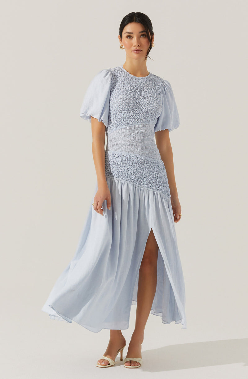A woman is wearing a long, light blue dress with a fitted bodice and a flowing skirt. The dress features puffed short sleeves made of sheer fabric, with gathered details. The bodice has textured, ruched fabric with smocking, and the skirt has a high slit revealing one leg. The woman is also wearing beige high-heeled shoes.