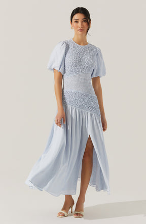 A woman is wearing a long, light blue dress with a fitted bodice and a flowing skirt. The dress features puffed short sleeves made of sheer fabric, with gathered details. The bodice has textured, ruched fabric with smocking, and the skirt has a high slit revealing one leg. The woman is also wearing beige high-heeled shoes.
