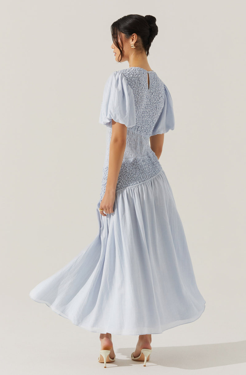 A woman is wearing a light blue dress with short, puffed sleeves that are gathered at the cuffs. The dress has a fitted, smocked bodice with a textured, gathered pattern and a flowy, floor-length skirt made of lightweight fabric. The back of the dress features a small keyhole opening near the neckline. She is also wearing beige high-heeled shoes.