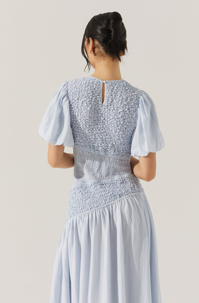 The image shows a woman wearing a light blue dress. The dress features a textured, ruched bodice with a small keyhole opening at the back, fastened with a button. The dress has voluminous puff sleeves and a high waistline with gathered fabric that extends into a flowing, ankle-length skirt.