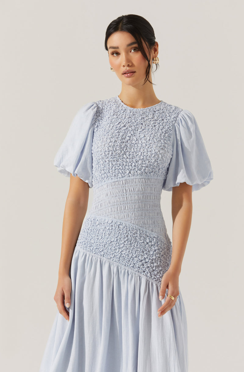 A woman is wearing a light blue dress with a fitted bodice and puff sleeves. The bodice features textured, smocking details with a mix of ruched and gathered fabric, creating a patterned appearance. The skirt is flowing and gathered, made from a lightweight, slightly crinkled fabric.