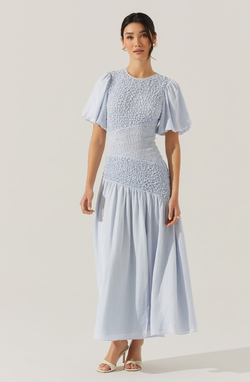 A woman is wearing a light blue dress with a fitted bodice featuring textured, gathered fabric. The dress has puffy short sleeves and a full-length skirt with a gathered, flowing design. She is also wearing open-toe heels.
