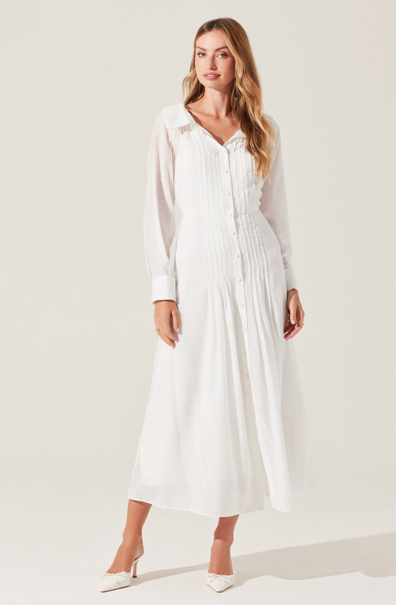 A woman is wearing a long, white, button-up dress with a collared neckline. The dress features vertical pleats and sheer, long sleeves with wide cuffs. She is also wearing white high-heeled shoes.