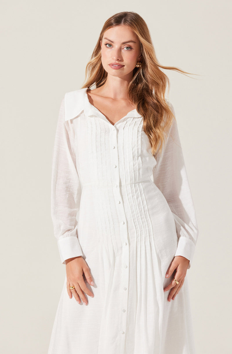 A woman is wearing a long, white dress with a button-down front and a collar. The dress features vertical pleats running down the front and long, semi-sheer sleeves with wide cuffs. The fabric appears lightweight and slightly textured.