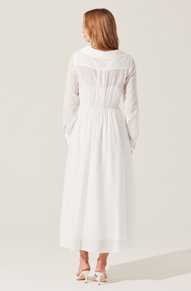 A woman is wearing a long, white, flowing dress with long sleeves. The dress has a fitted waist and a gathered skirt that reaches her ankles. The fabric appears lightweight and semi-sheer, giving it an airy, elegant look. She is also wearing a pair of white heeled sandals.