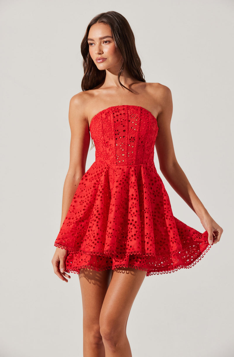 A woman is wearing a strapless, red dress with eyelet embroidery. The dress has a fitted bodice and a flared, layered skirt that falls above the knees. The eyelet pattern creates a textured, decorative look throughout the dress.