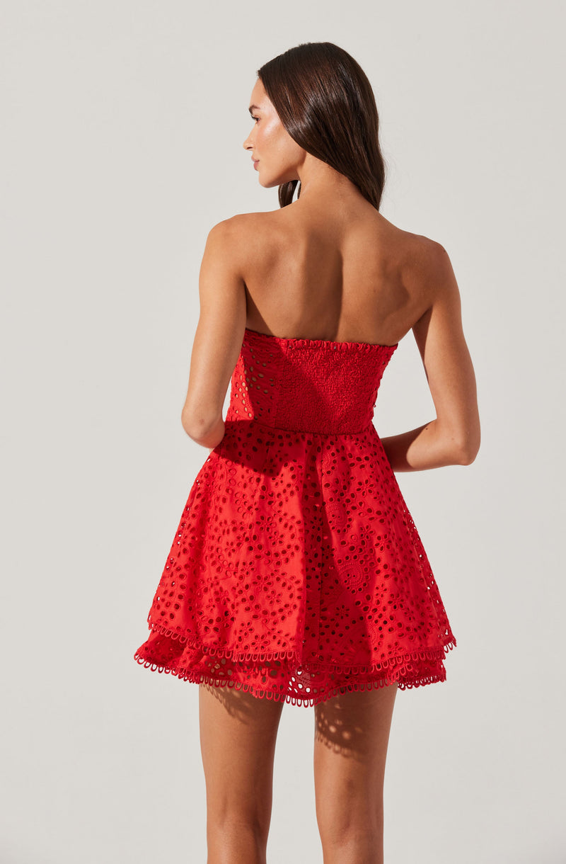 A woman is wearing a strapless, red dress with a fitted bodice and a flared skirt. The fabric features a pattern of small, circular cutouts, creating a semi-transparent look. The hemline is decorated with a scalloped lace trim.