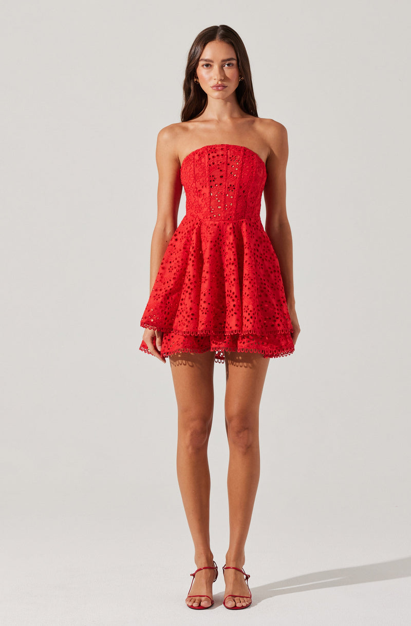 A woman is wearing a strapless, bright red dress with eyelet embroidery. The dress features a fitted bodice with vertical paneling and a flared, layered skirt that falls above the knee. She is also wearing matching red open-toe high-heel sandals with ankle straps.