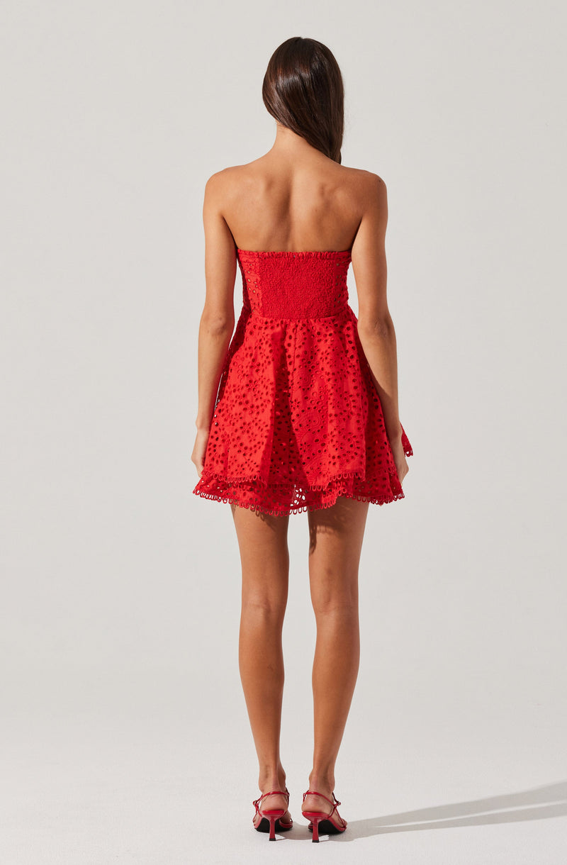 A woman is wearing a strapless red dress with eyelet embroidery and a gathered, smocked bodice. The skirt is short, flared, and decorated with circular cutouts and eyelet detailing at the hem. She is also wearing red high-heeled sandals with crossed straps.