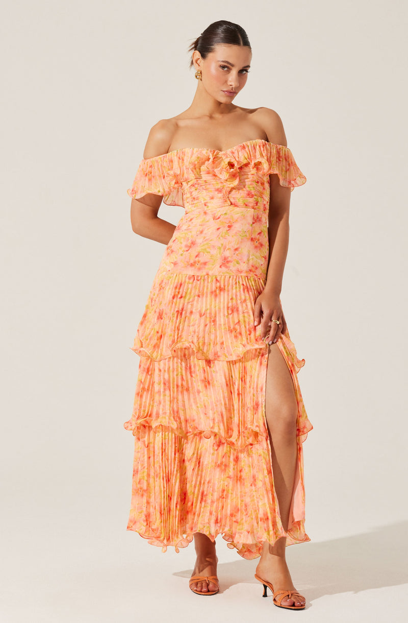 A woman is wearing an off-the-shoulder maxi dress with ruffles along the neckline and tiered, ruffled layers down the length of the skirt. The dress features a tie-dye pattern in shades of pink and orange, with a high slit on one side revealing her leg.