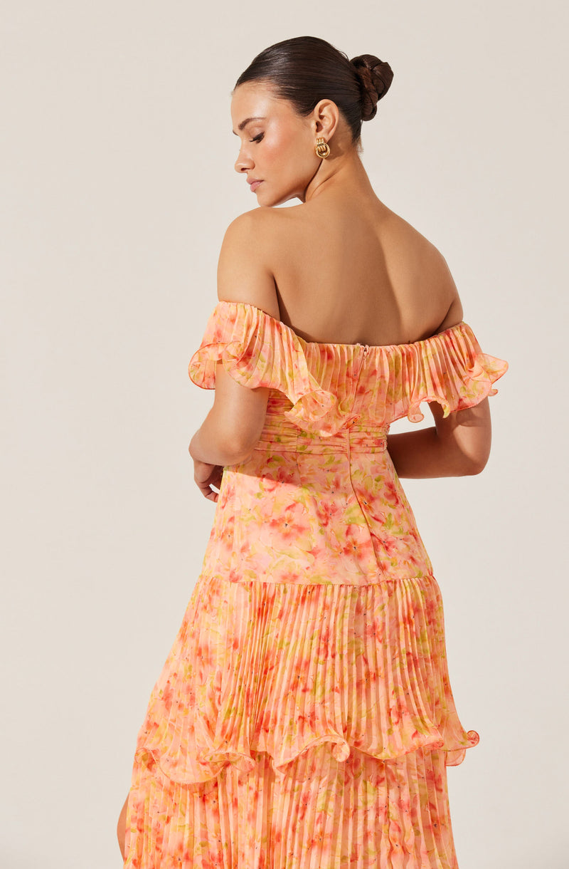 A woman is wearing an off-the-shoulder dress with ruffled sleeves. The dress features a blend of soft pastel colors, including shades of pink, orange, and yellow, with a pattern that appears to be a mix of tie-dye and vertical stripes. The fabric looks lightweight and flowy, and the dress has a tiered design with ruffles and pleats. She is accessorized with gold hoop earrings and has her dark hair styled in a sleek, low bun.