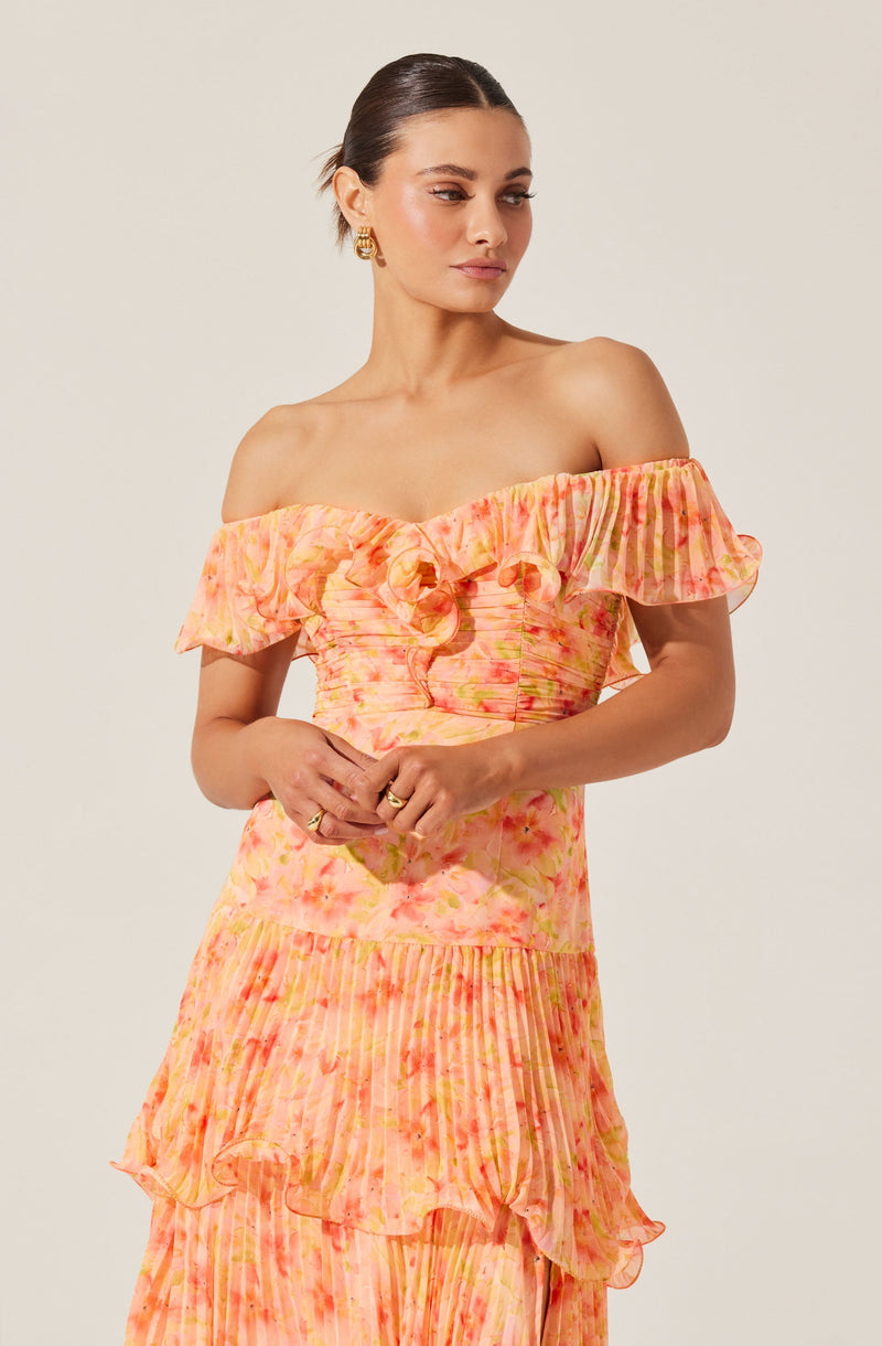A woman is wearing a dress with an off-the-shoulder neckline and short ruffled sleeves. The dress features a soft, pastel watercolor pattern in shades of pink, orange, and yellow, with a smocked bodice and layered, ruffled skirt sections.
