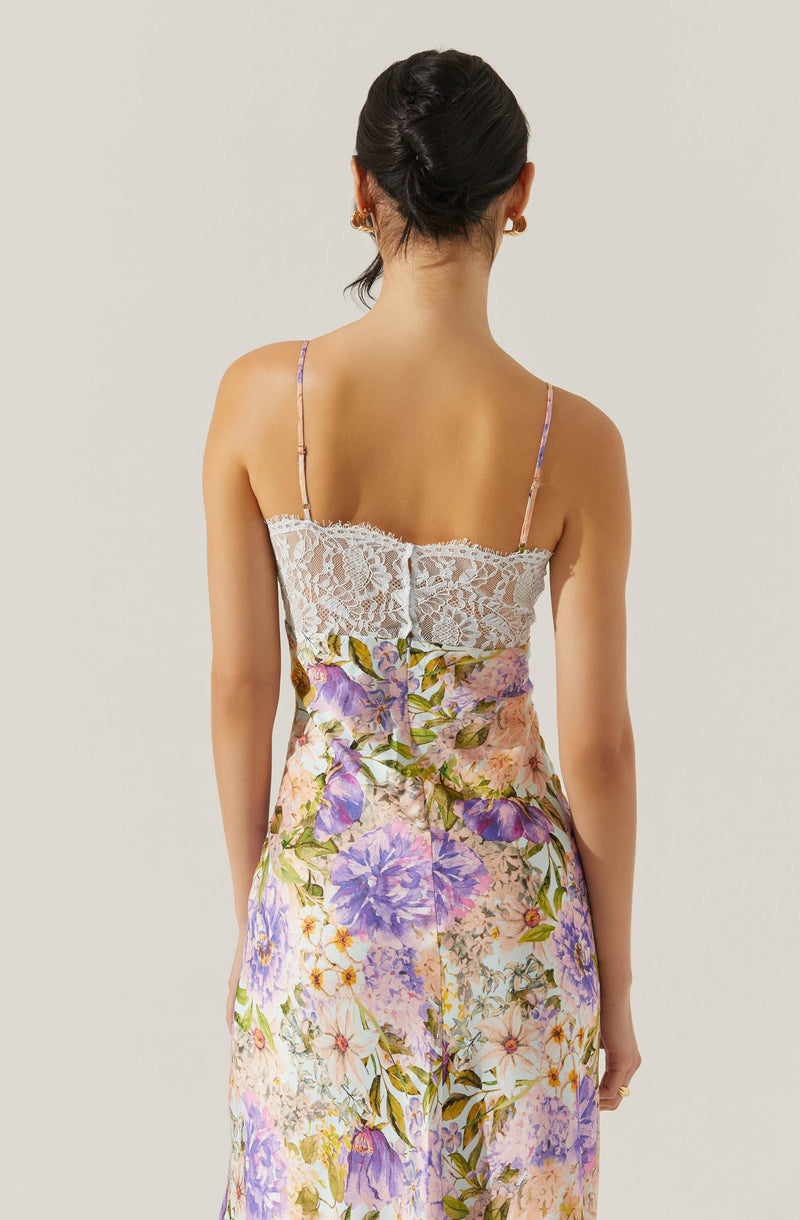 A woman is wearing a dress with a lace bodice and floral print fabric. The dress has thin, delicate spaghetti straps. The floral pattern features shades of purple, pink, green, and white, creating a soft, feminine look.