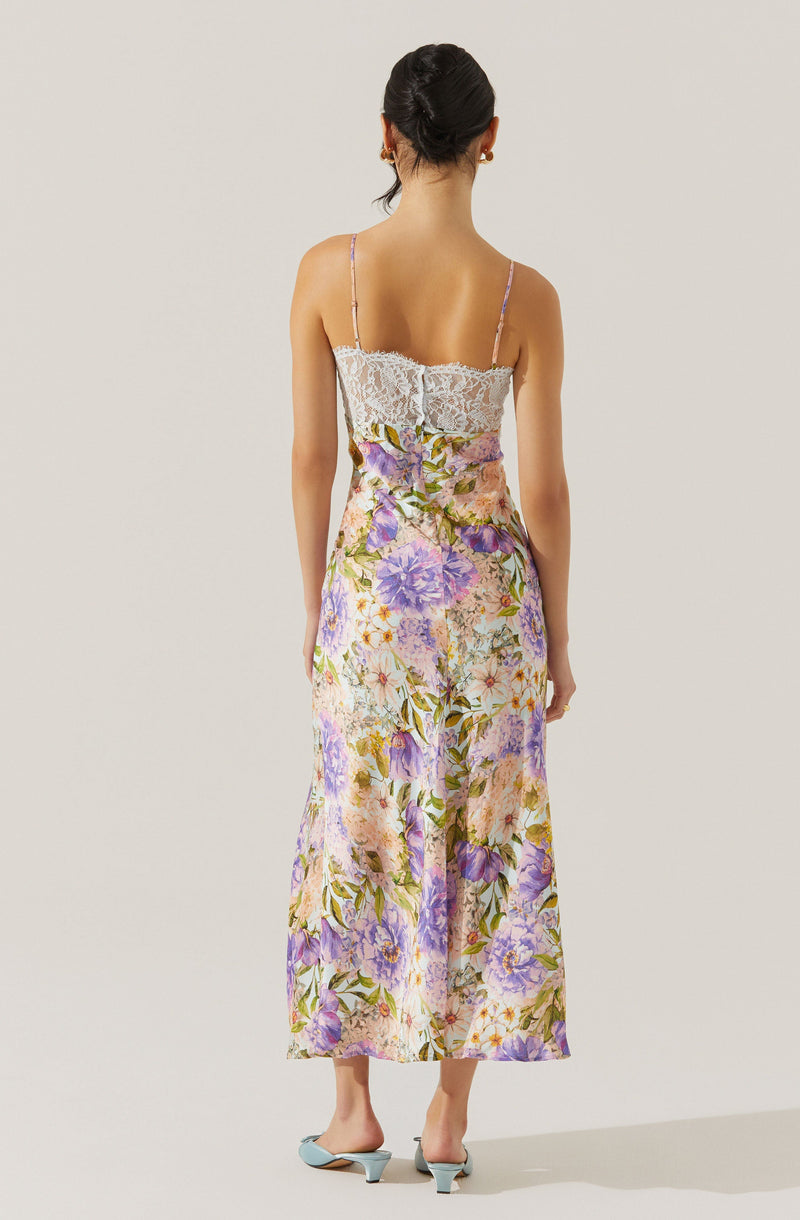 The dress features a fitted bodice with white lace fabric and thin spaghetti straps. The skirt is long, flowing, and has a vibrant floral print with shades of purple, pink, green, and yellow on a light background. The dress reaches down to the ankles, and the wearer is also wearing light-colored heeled shoes.