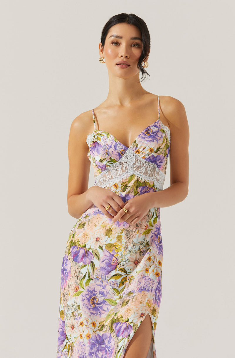 A woman is wearing a sleeveless, floral dress with spaghetti straps. The dress features a vibrant pattern of purple, lavender, pink, and yellow flowers with green leaves on a light background. The top of the dress has a lace detail under the bust, adding a delicate touch. The skirt has a slit on one side, showing a bit of leg.
