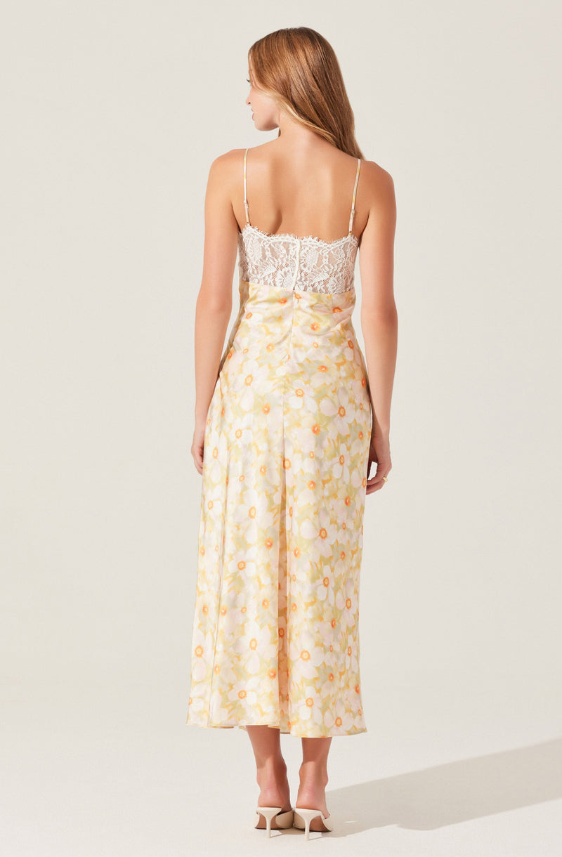 A woman is wearing a sleeveless, fitted top made of white lace fabric with delicate spaghetti straps. She is also wearing high-waisted, wide-leg pants with a light yellow base color and a floral pattern featuring orange and yellow flowers.