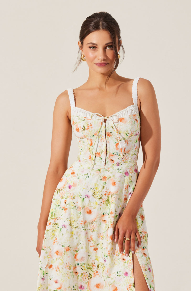 A woman is wearing a sleeveless, white dress with a floral pattern in shades of orange, pink, green, and yellow. The dress features wide straps, a gathered neckline with a small tie at the center, and a side slit.