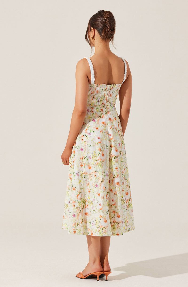 A woman is wearing a sleeveless, knee-length dress with a fitted bodice and a full skirt. The dress features wide straps with white lace trim and is made of a light, floral-printed fabric with pastel colors, including pink, orange, green, and white. The overall style is feminine and summery. The woman is also wearing open-toed high heels and has her hair styled in an elegant updo.