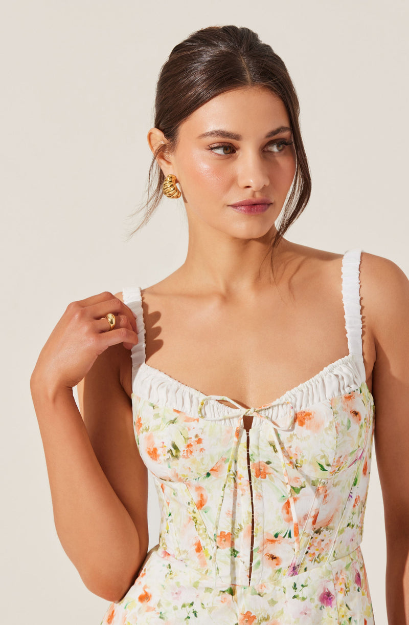 A woman is wearing a sleeveless, white dress with ruffled straps and a floral print in shades of orange, pink, and green. The dress has a gathered neckline with a small bow and a front zipper.