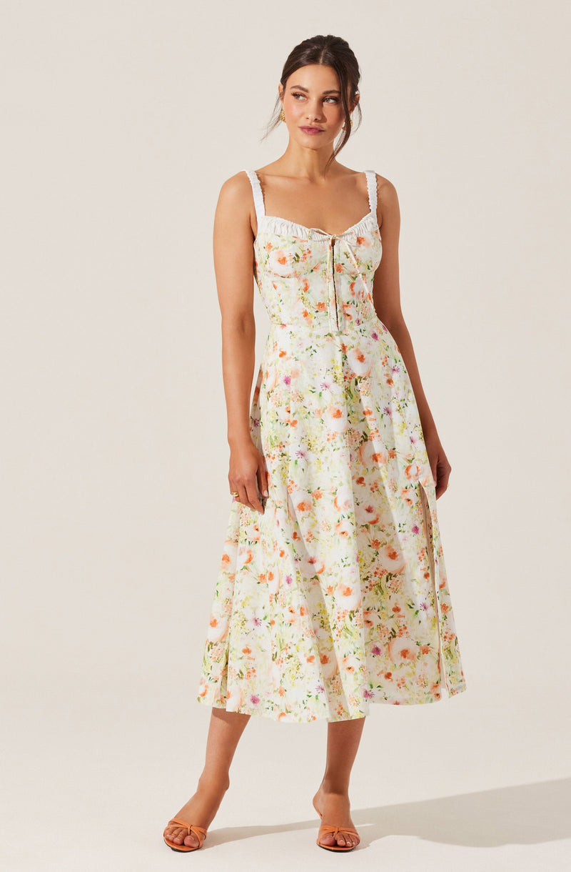 A woman is wearing a sleeveless, ankle-length dress with a floral pattern in pastel colors. The dress has wide, textured straps and a gathered neckline with a small bow at the center. The fabric features a light background with a variety of soft, colorful flowers. She is also wearing tan, open-toe heels.